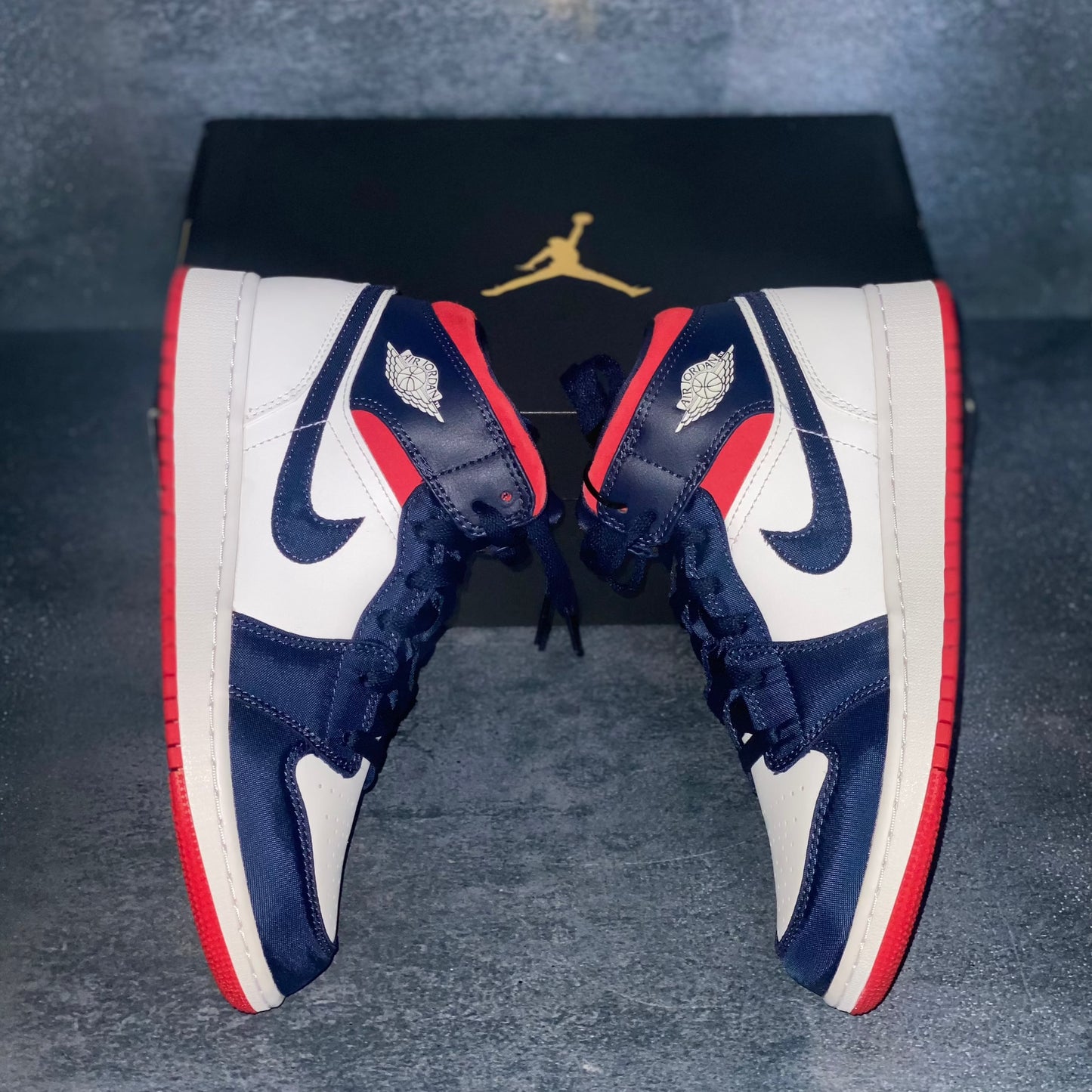 The lateral side of the red, white, and blue Nike Air Jordan 1 Mid sneakers with a black Air Jordan sneaker box.