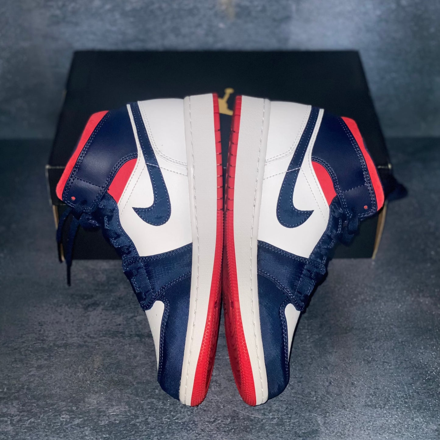 The medial side of the red, white, and blue Nike Air Jordan 1 Mid sneakers with a black Air Jordan sneaker box.