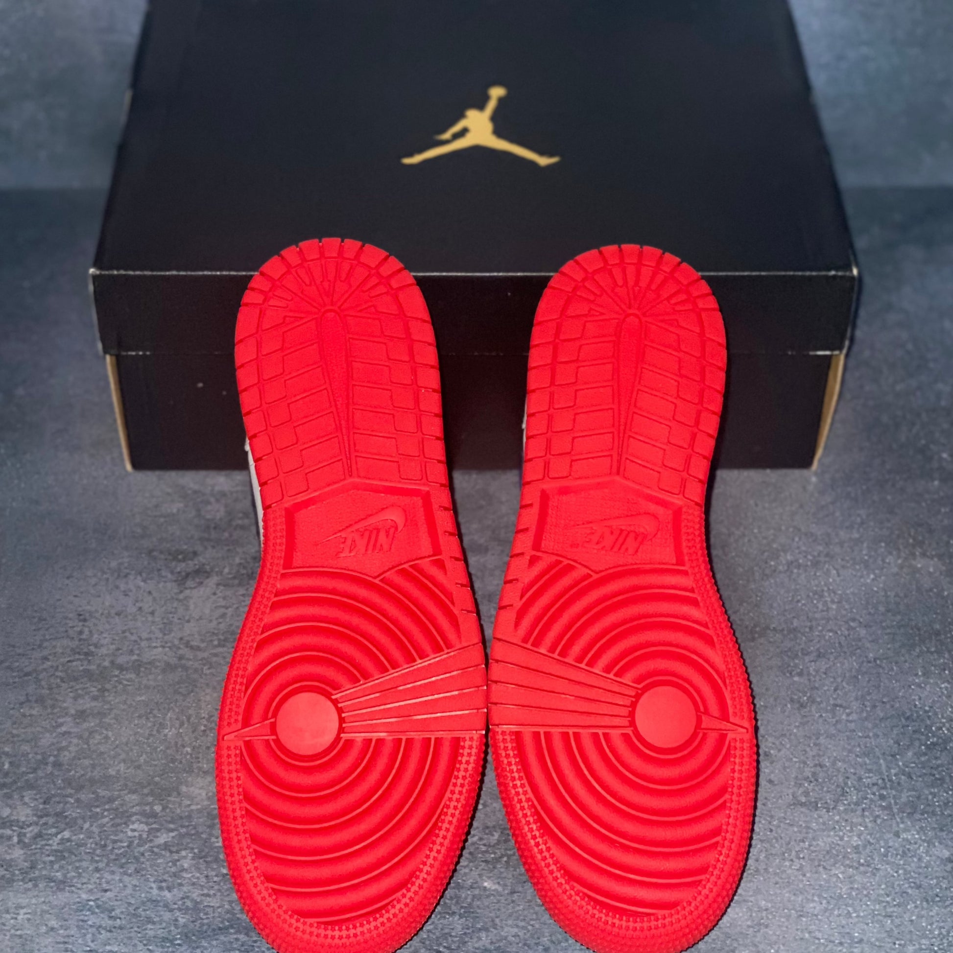 The red sneaker bottom of the red, white, and blue Nike Air Jordan 1 Mid sneakers with a black Air Jordan sneaker box.