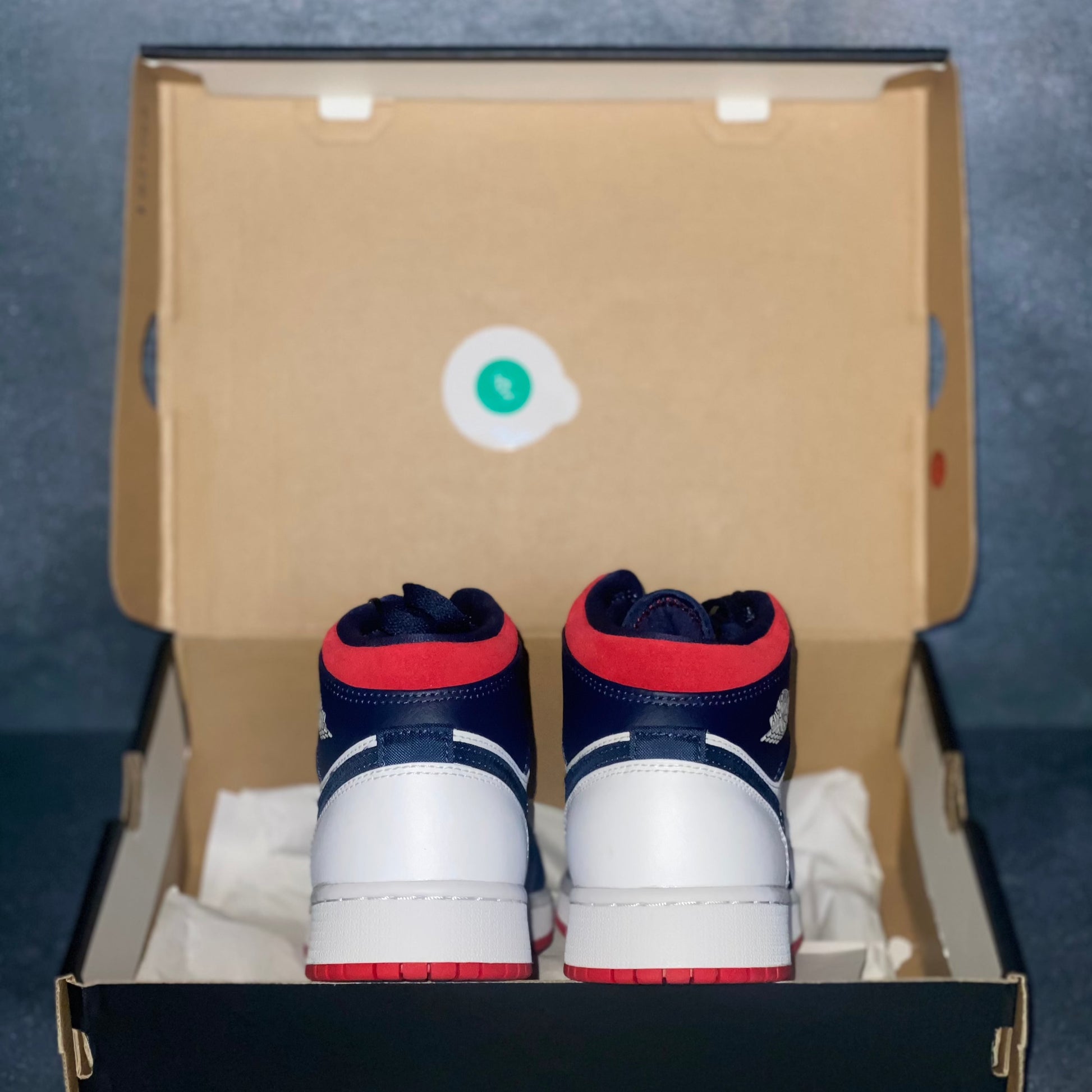 The heels of the red, white, and blue Nike Air Jordan 1 Mid sneakers with a black Air Jordan sneaker box.