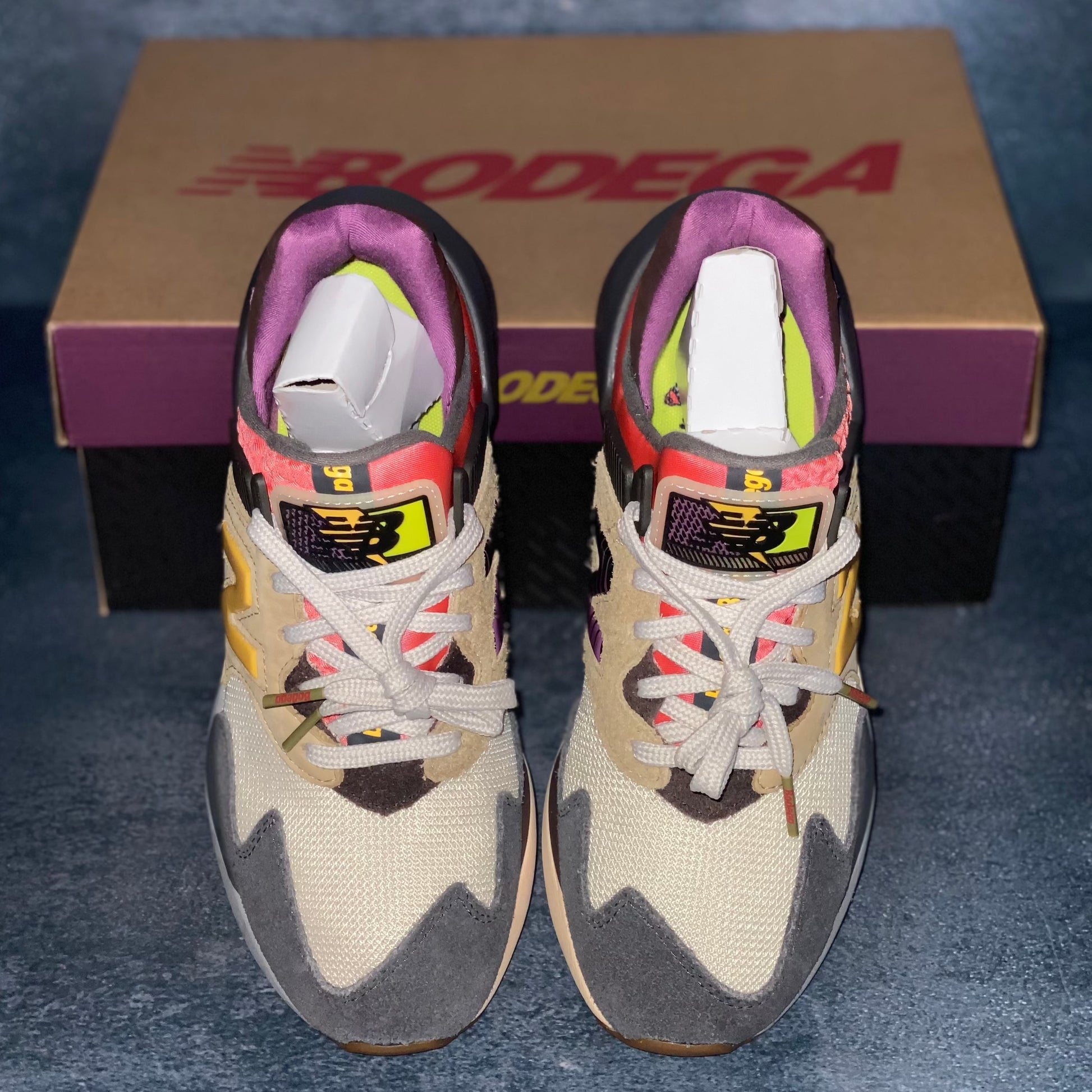 Beige, grey, and red multicolored New Balance 997S sneakers with a brown, purple, and black New Balance Bodega sneaker box.