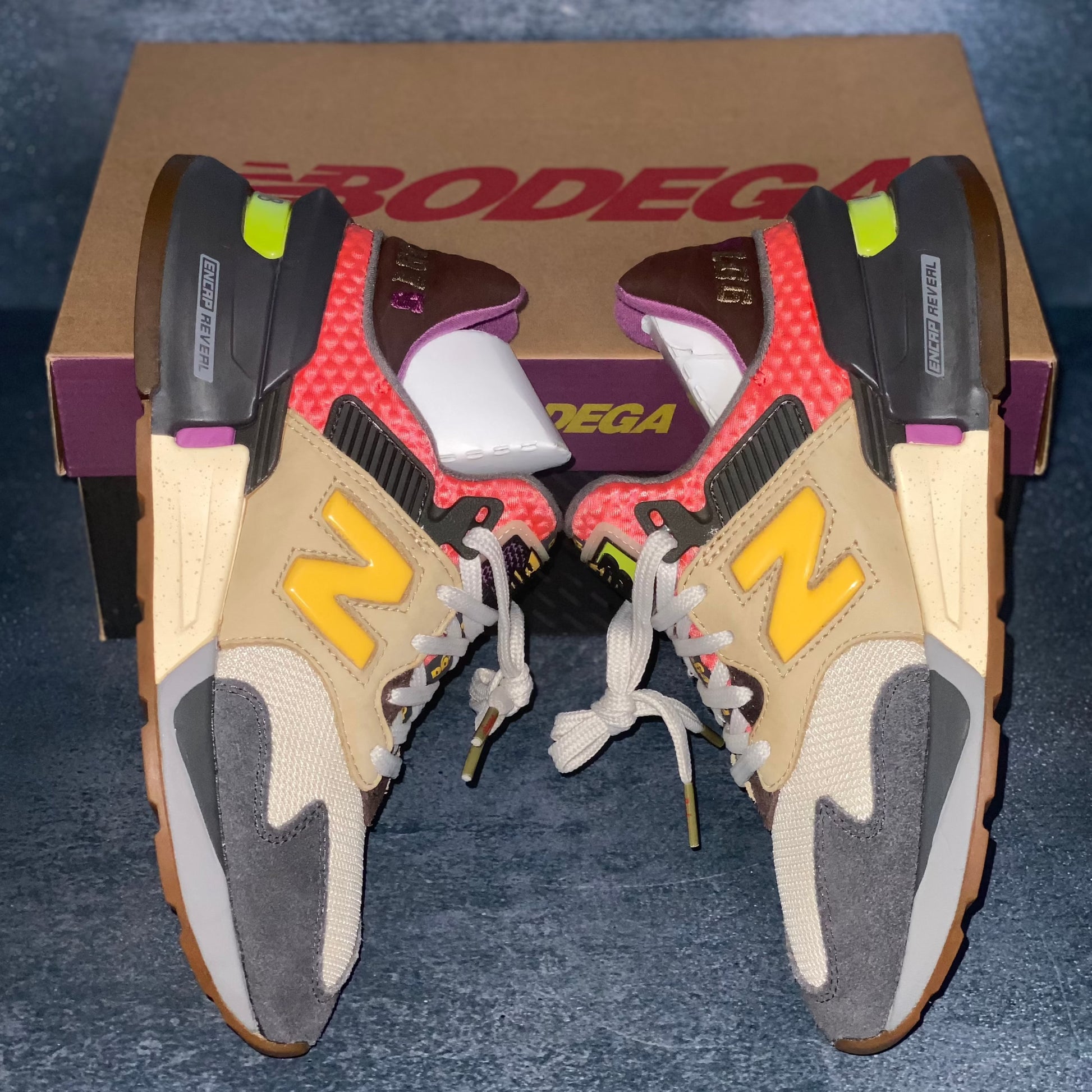 The lateral side of the beige, grey, and red multicolored New Balance 997S sneakers with a brown, purple, and black New Balance Bodega sneaker box.