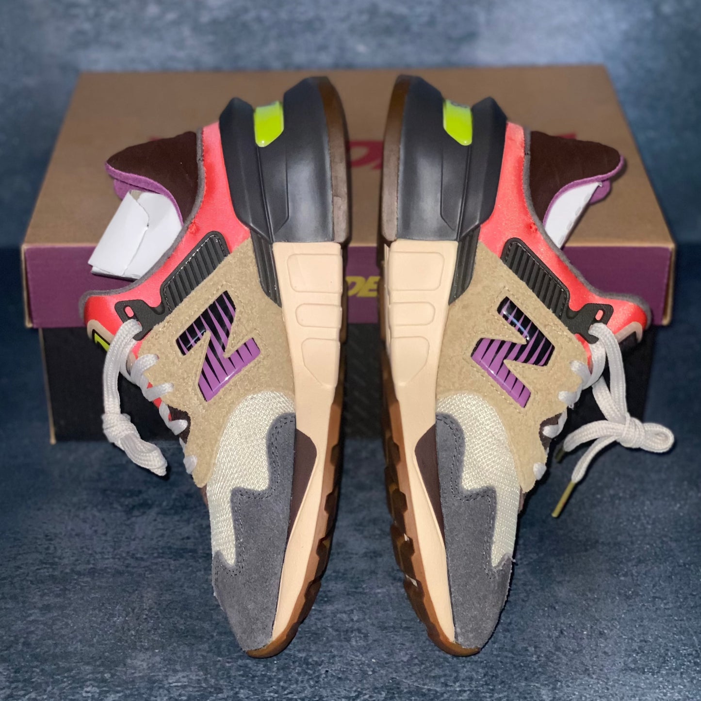 The medial side of the beige, grey, and red multicolored New Balance 997S sneakers with a brown, purple, and black New Balance Bodega sneaker box.