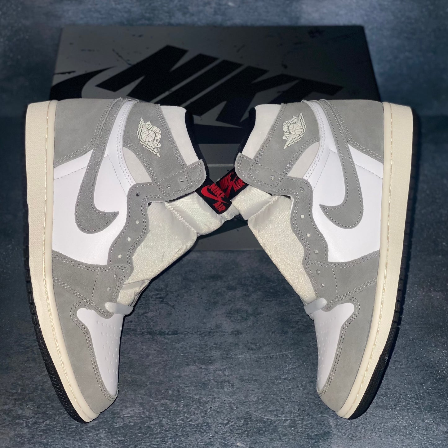 The lateral side of the white and grey Nike Air Jordan 1 Retro High sneakers with a black and grey Nike Air Jordan sneaker box.