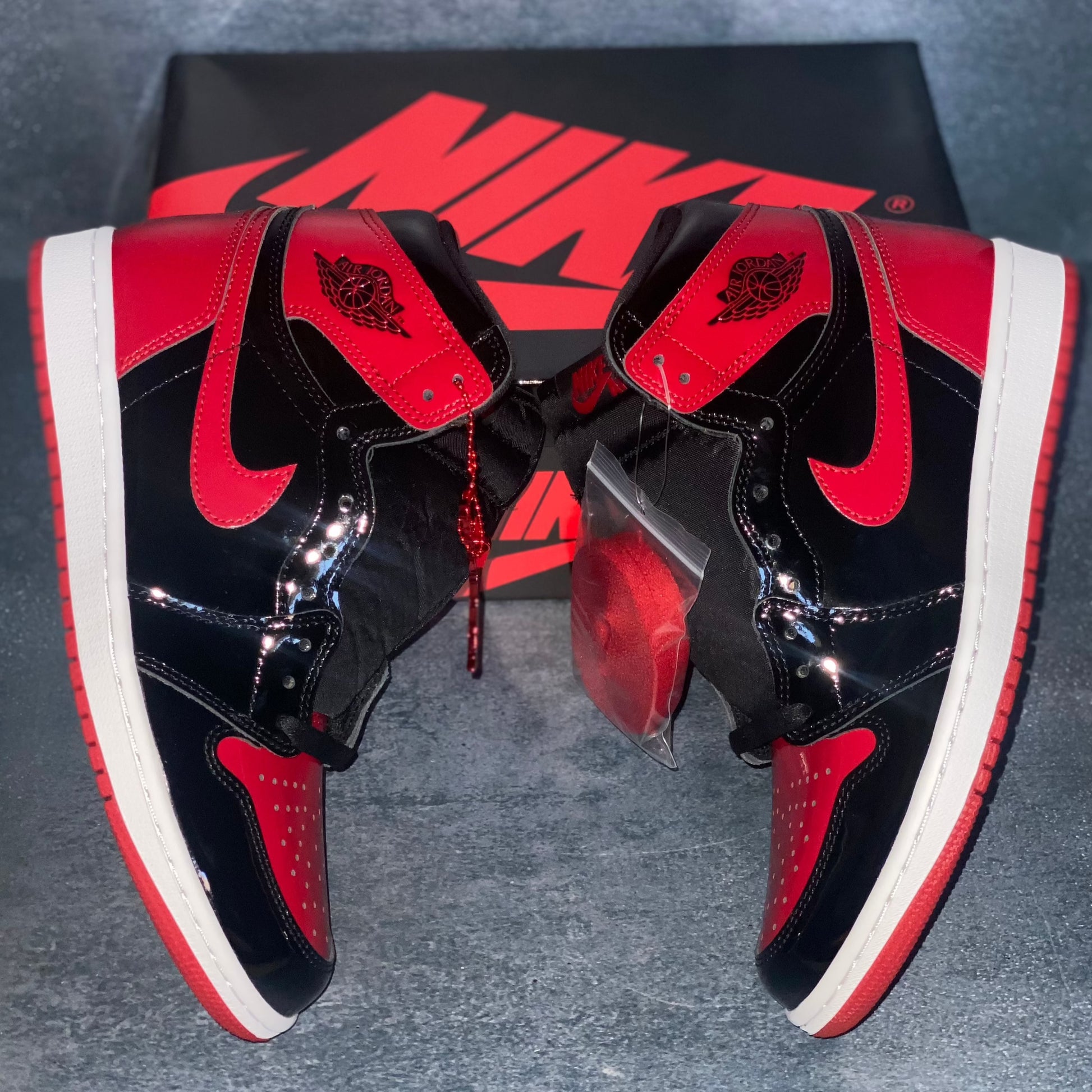 The lateral side of the black and red patent leather Nike Air Jordan 1 Retro High sneakers with a black and red Nike Air Jordan sneaker box.